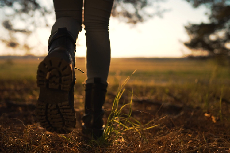Kuvassa näkyy retkeilijän jalat maastossa kävelemässä kohti aurinkoista maisemaa.